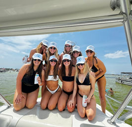 Group of nine women in bikinis and matching white caps smiling and holding canned drinks on the bow of a boat during a sunny summer boat party in a busy coastal bay