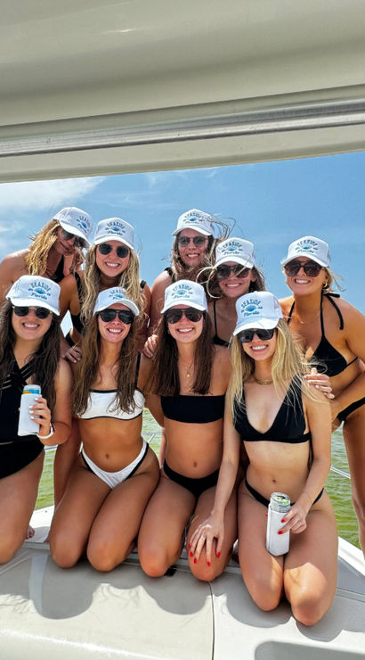 Group of nine women in bikinis and matching white caps smiling and holding canned drinks on the bow of a boat during a sunny summer boat party in a busy coastal bay
