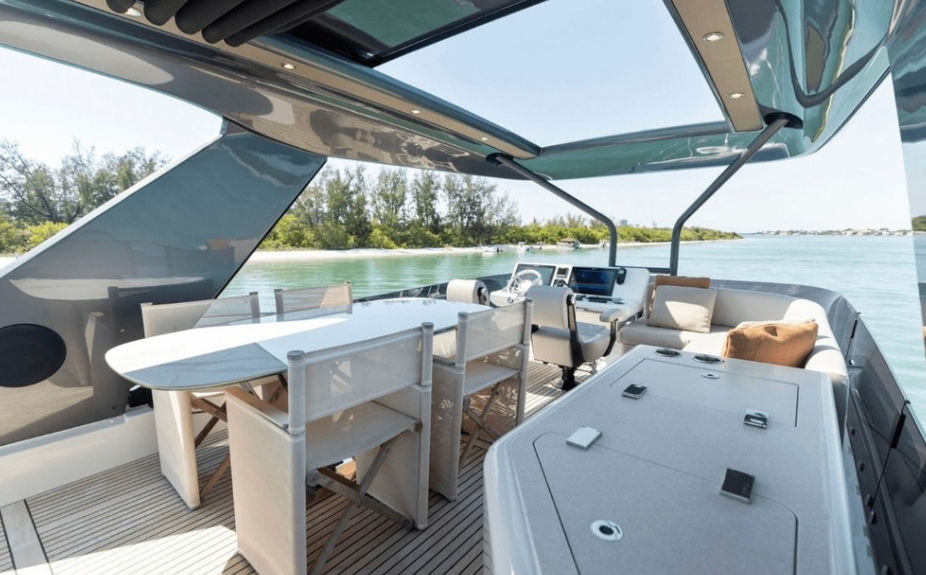 Luxury yacht aft deck with shaded outdoor dining table, cushioned lounge seating and helm station overlooking turquoise tropical inlet and tree-lined shoreline