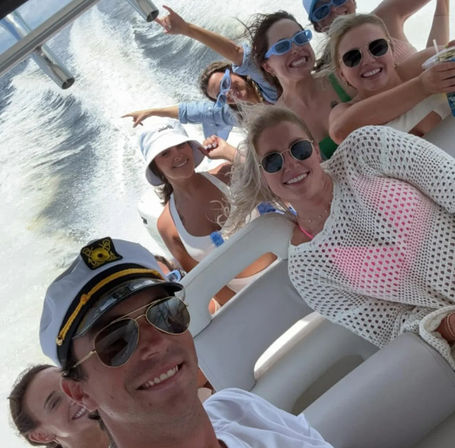 Group of friends smiling on a speedboat ride — captain's hat in the foreground, sunglasses and swimwear, white wake trailing across sunlit coastal waters, summer boat party vibe