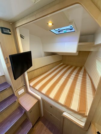 Cozy yacht cabin with striped orange-and-white mattress, built-in wood drawers and shelving, wall-mounted TV, small steps leading down, and overhead hatch skylight