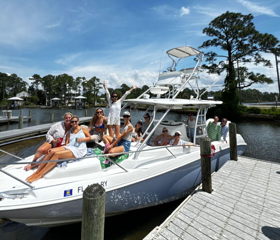 Group of people lounging on a white motorboat tied to a wooden dock at a sunny Florida marina, pine trees and blue sky overhead.