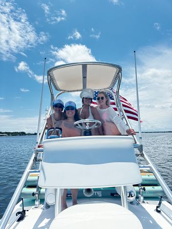 Four friends smiling on a motorboat under a white T-top with an American flag, wearing sunglasses and hats, cruising calm coastal waters beneath a bright blue summer sky.