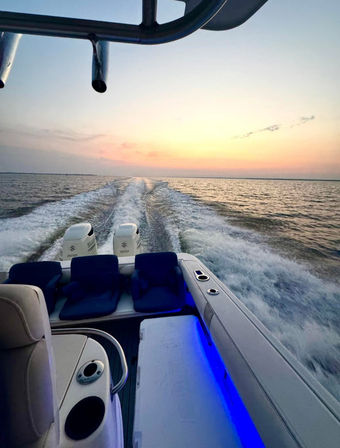 Dusk boat ride on open water with twin outboard engines carving a foamy wake toward a pink-orange sunset; blue LED-lit deck and cushioned seats on a powerboat.