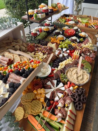Colorful outdoor grazing table on wooden boards with charcuterie: rolled cured meats, assorted cheeses, crackers, berries, grapes, sliced fruit, veggie sticks, dips and mini desserts for a garden party.