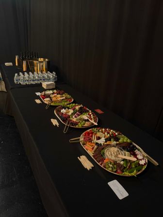 Indoor event catering setup: long black buffet table with colorful charcuterie platters of cheeses, cured meats, fruits, crackers and dip, plus bottled water, wine buckets and napkins.