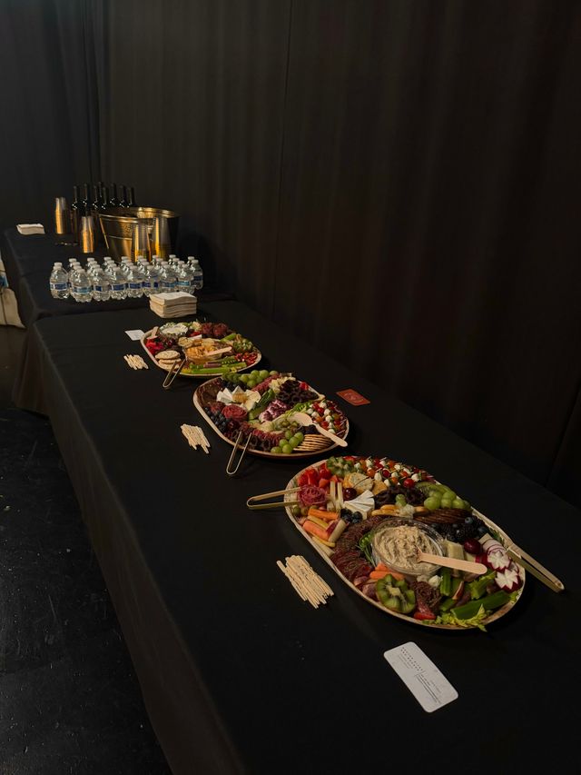 Indoor event catering setup: long black buffet table with colorful charcuterie platters of cheeses, cured meats, fruits, crackers and dip, plus bottled water, wine buckets and napkins.