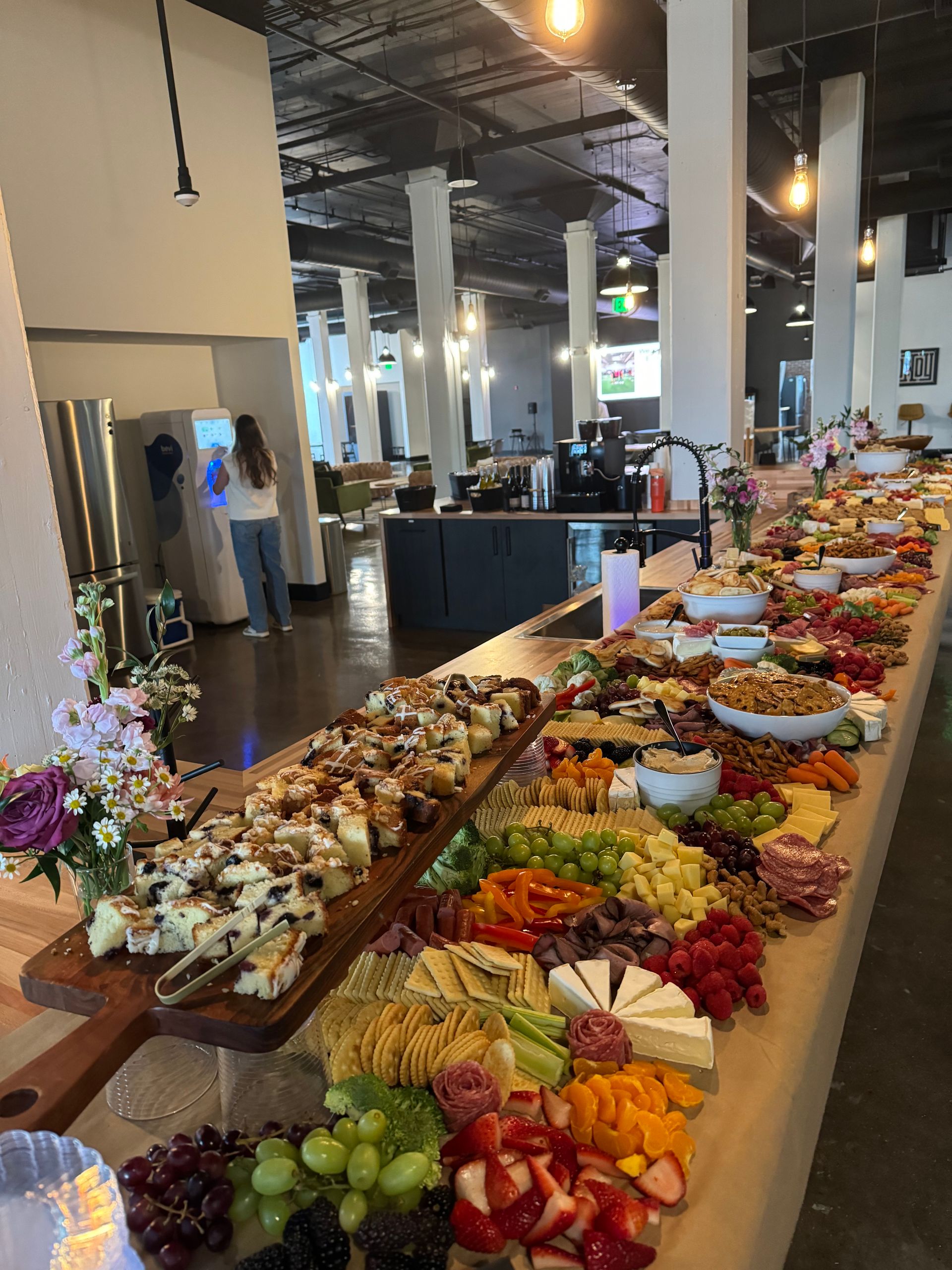 Mouthwatering charcuterie and dessert buffet with cheeses, fruits, crackers and pastries on a long wooden table in a modern industrial loft event space with pendant lights and a person at a drink station in the background.
