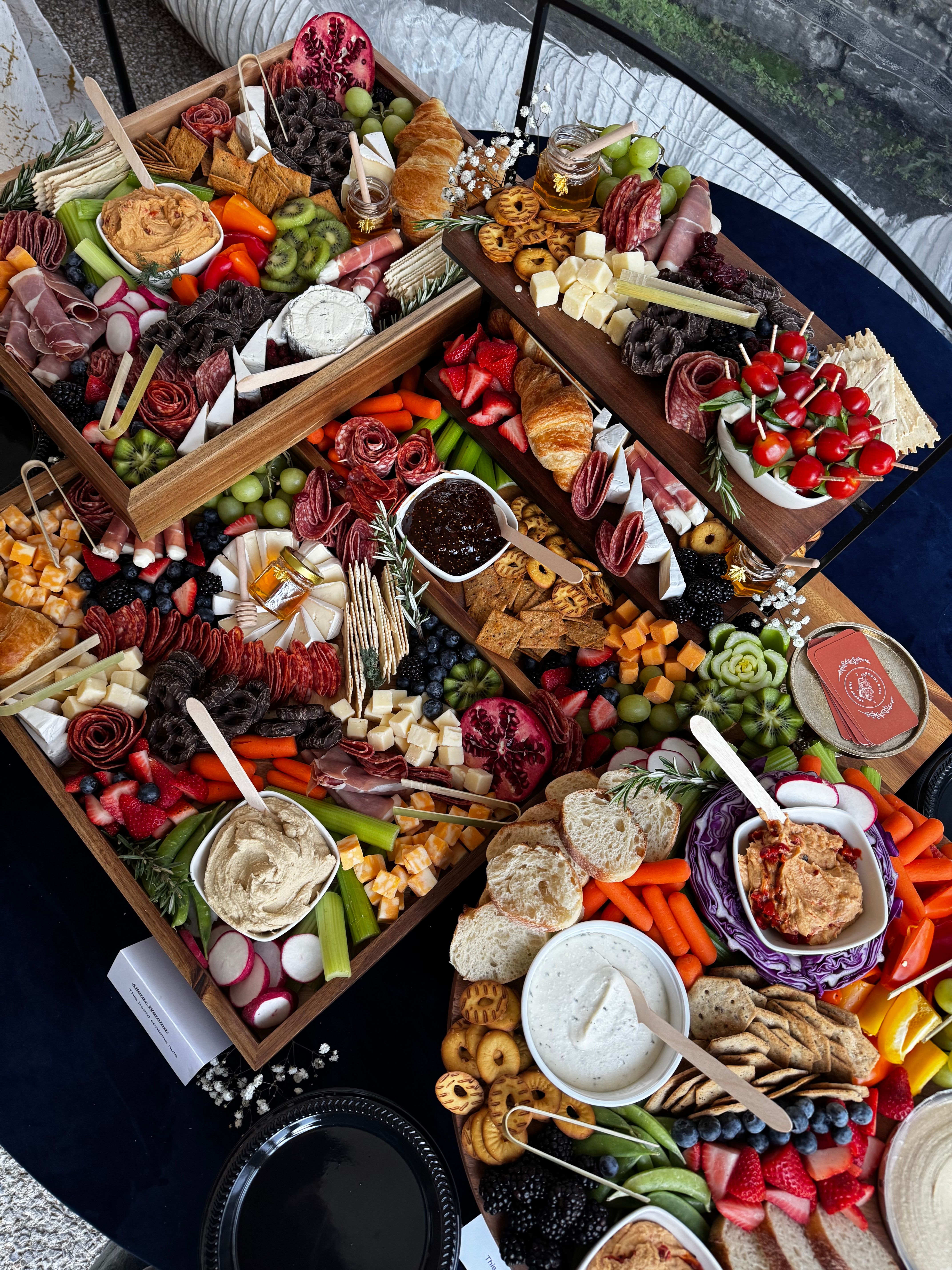 Vibrant party charcuterie spread on wooden boards with assorted cured meats, cheese cubes and wedges, croissants, crackers, skewered cherry tomatoes, hummus and creamy dips, fresh fruit (strawberries, grapes, blueberries, kiwi, pomegranate), veggies and pretzels—stylish grazing board for events and gatherings.