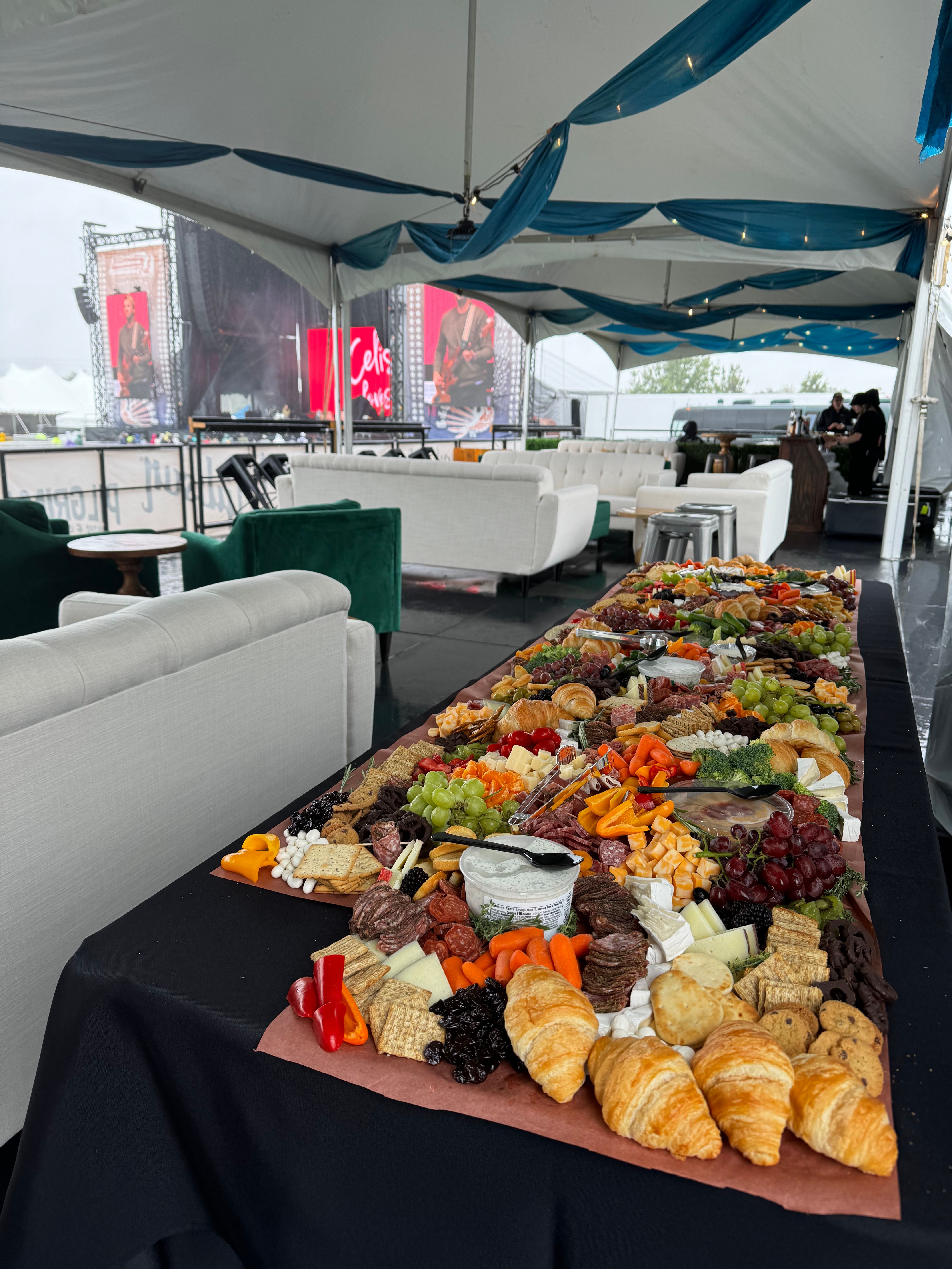 Mouthwatering charcuterie grazing table with croissants, cheeses, fruits, crackers and cured meats set on a long table inside an outdoor festival VIP tent with white lounge sofas and concert stage screens visible in the background.