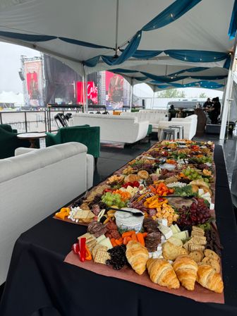 Mouthwatering charcuterie grazing table with croissants, cheeses, fruits, crackers and cured meats set on a long table inside an outdoor festival VIP tent with white lounge sofas and concert stage screens visible in the background.