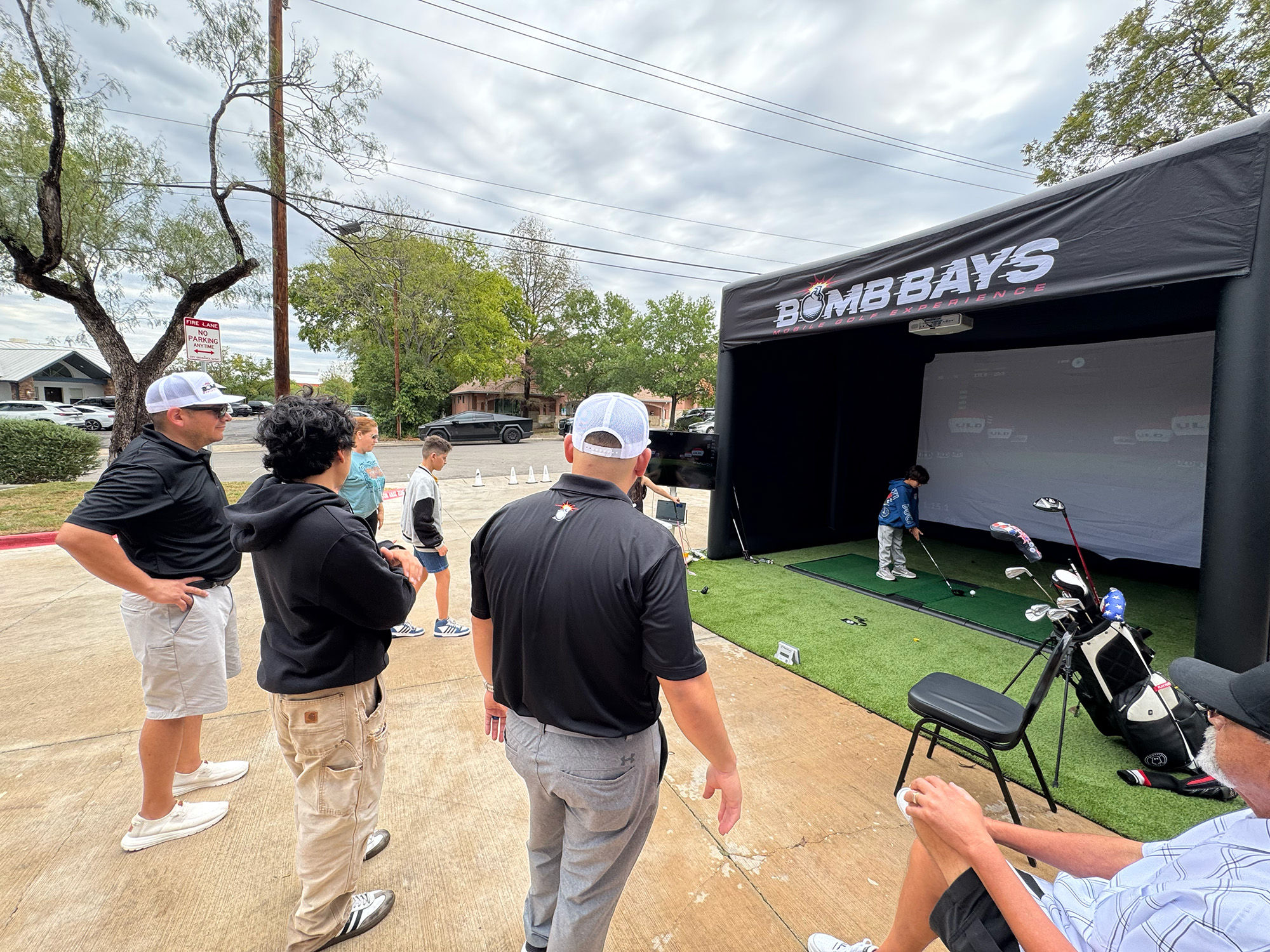 Small crowd watches a child swing golf clubs inside a black inflatable golf simulator booth on artificial turf at an outdoor suburban parking-lot demo