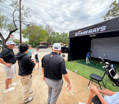 Small crowd watches a child swing golf clubs inside a black inflatable golf simulator booth on artificial turf at an outdoor suburban parking-lot demo