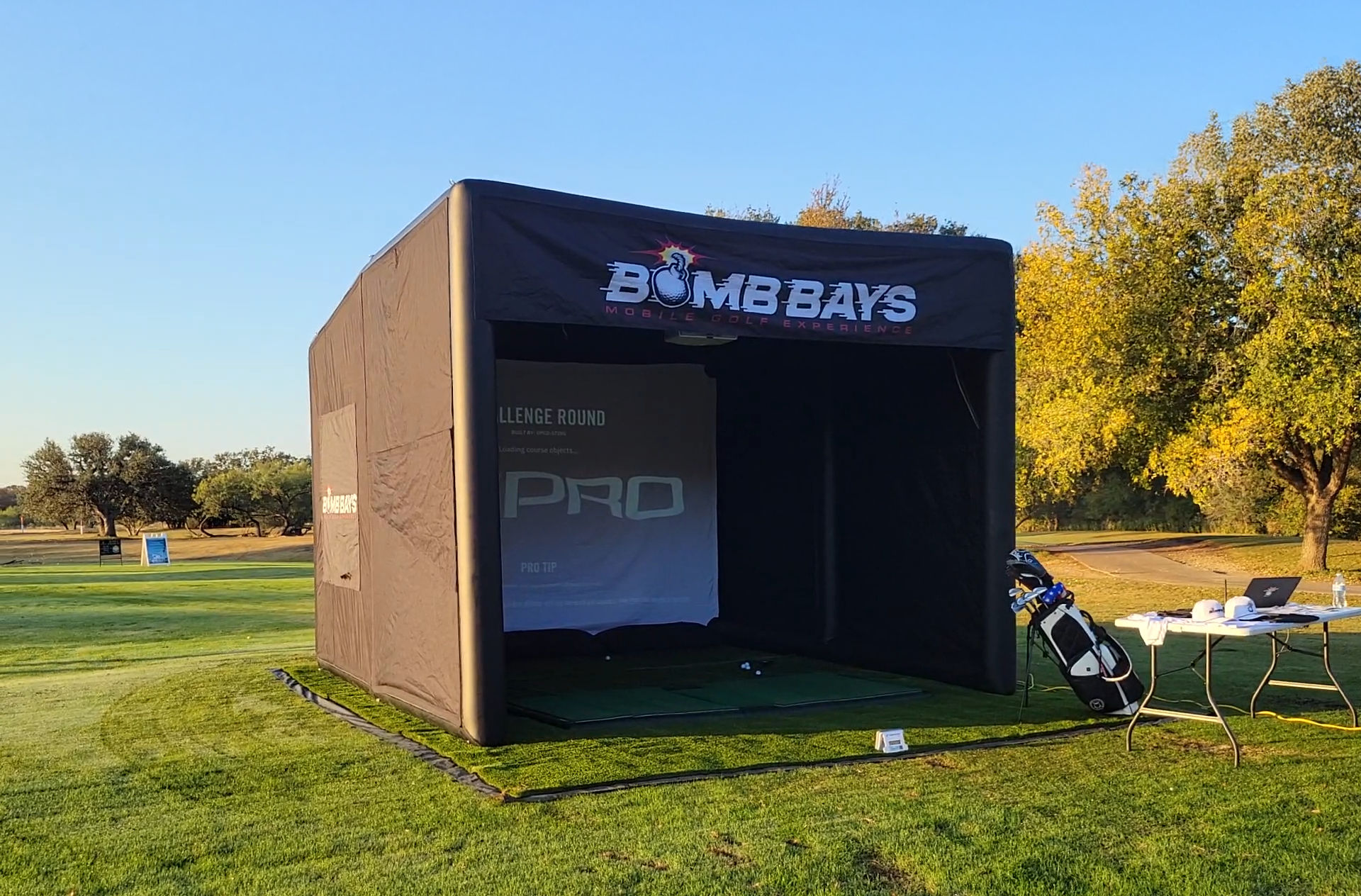 Outdoor portable golf simulator bay on a sunny driving range — black inflatable practice tent on green turf with a golf bag and folding table of gear, trees and clear blue sky in the background