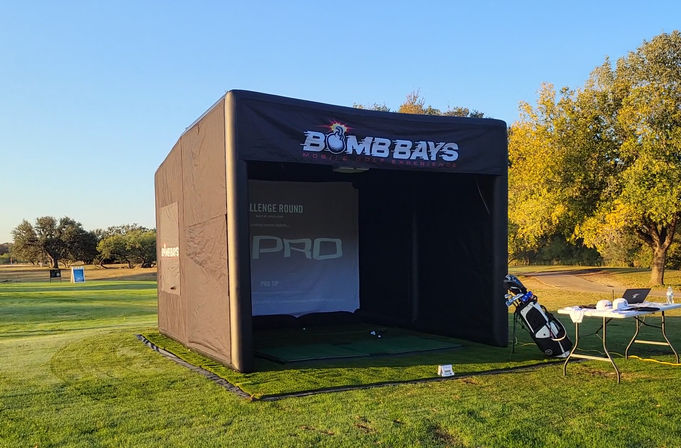Outdoor portable golf simulator bay on a sunny driving range — black inflatable practice tent on green turf with a golf bag and folding table of gear, trees and clear blue sky in the background