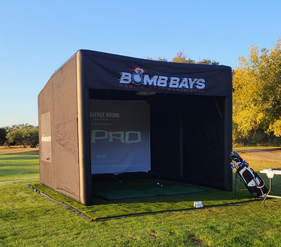 Outdoor portable golf simulator bay on a sunny driving range — black inflatable practice tent on green turf with a golf bag and folding table of gear, trees and clear blue sky in the background