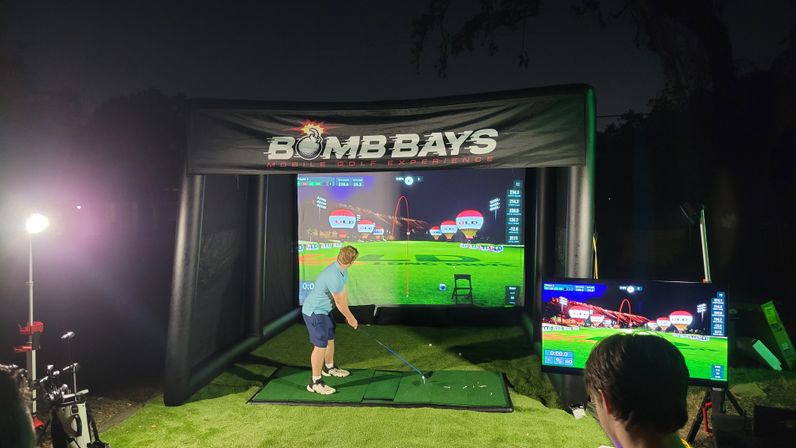Person mid-swing in an inflatable outdoor golf simulator at night, hitting off artificial turf toward a large virtual driving-range screen and side monitor.