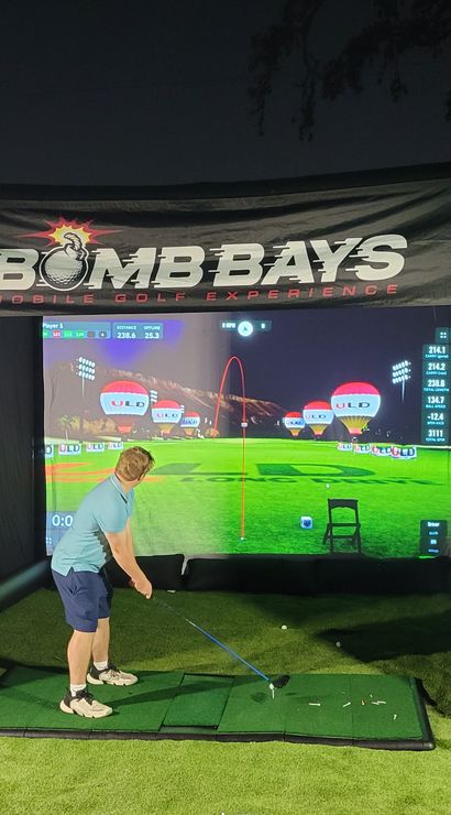 Person mid-swing in an inflatable outdoor golf simulator at night, hitting off artificial turf toward a large virtual driving-range screen and side monitor.