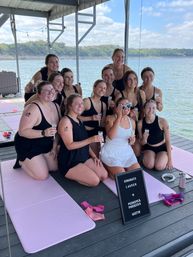 Group of women in black swimsuits on a lakeside dock under a boathouse, seated on pink mats and raising prosecco flutes around a guest in white with heart-shaped sunglasses and a congratulatory letter board.