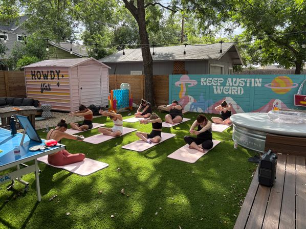 Group of yoga participants doing seated stretches on pink mats in a shaded Austin backyard with a colorful "Keep Austin Weird" mural, a pink "Howdy y'all" shed, string lights and a round hot tub.