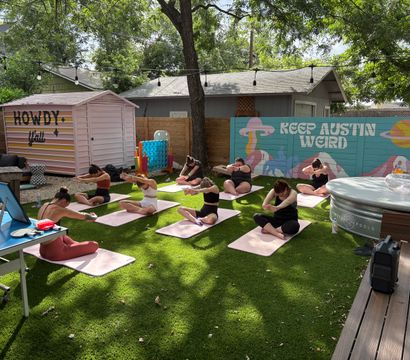Group of yoga participants doing seated stretches on pink mats in a shaded Austin backyard with a colorful "Keep Austin Weird" mural, a pink "Howdy y'all" shed, string lights and a round hot tub.