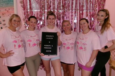 Six women in matching pink shirts smiling and holding a congratulatory letter board in front of pink streamers during a festive Austin celebration.