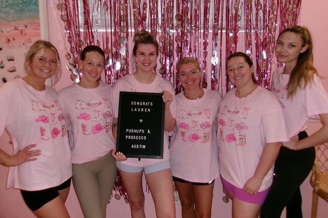Six women in matching pink shirts smiling and holding a congratulatory letter board in front of pink streamers during a festive Austin celebration.