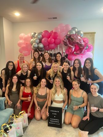 Smiling group of women posing at an indoor celebration with a pink and silver balloon garland, shimmering backdrop, champagne flutes and gift bags, holding a letter board congratulating the honoree.