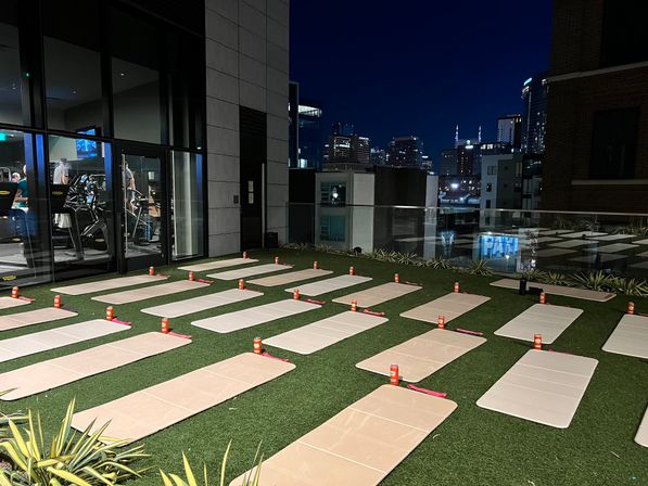 Moonlit urban rooftop fitness setup with rows of yoga mats and orange water bottles on artificial turf, glass-walled gym to the left and lit city skyline in the background.