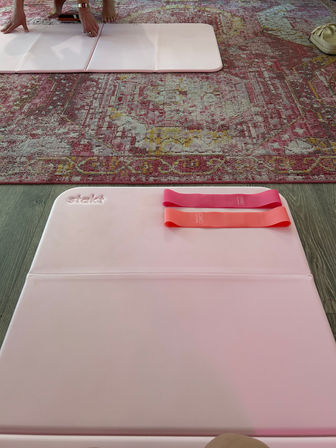 Living-room home workout scene with pink foldable yoga mats on wood floor, two pink/coral resistance bands, patterned area rug and bare feet in the background.