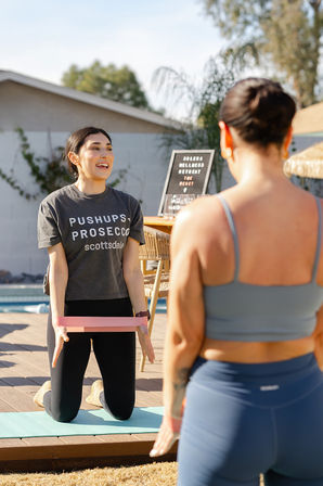 Sunlit poolside fitness class in Scottsdale: a kneeling instructor on a yoga mat holds a pink resistance band and smiles while guiding a standing participant in activewear, with a wellness retreat sign in the background.