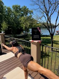Person in a brown workout set balancing on one leg for an outdoor workout on a sunny lakeside deck, yoga mat and a small congratulatory sign nearby with trees and calm water in the background.