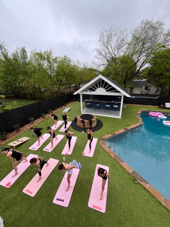 Aerial view of an outdoor yoga class on pink mats in a suburban backyard beside a swimming pool and white poolside pavilion under an overcast sky.