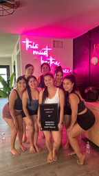 Group of seven women in athletic wear posing barefoot in a Scottsdale fitness studio with pink neon wall sign and plants, one holding a small congratulatory letter board.