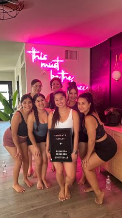 Group of seven women in athletic wear posing barefoot in a Scottsdale fitness studio with pink neon wall sign and plants, one holding a small congratulatory letter board.
