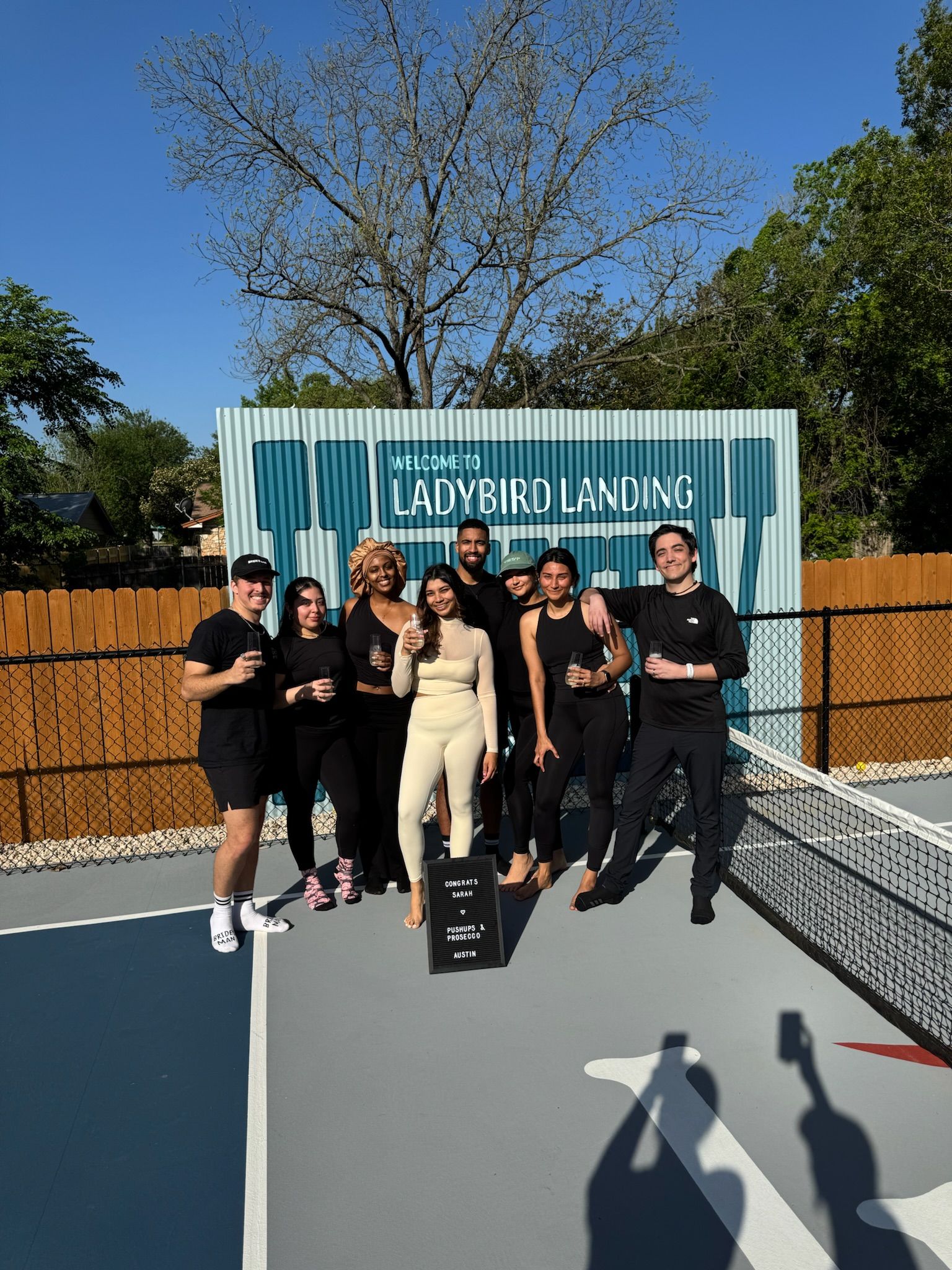 Eight friends smiling and holding drinks on a sunny outdoor pickleball court in Austin, Texas, posing in front of a blue welcome sign with trees and a wooden fence behind them.