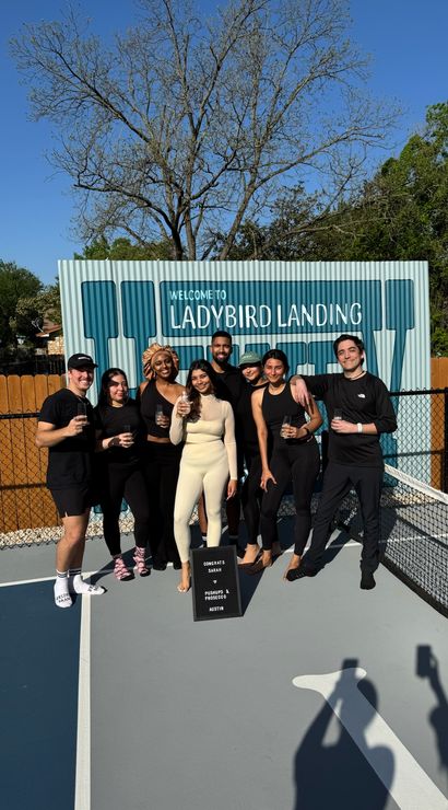 Eight friends smiling and holding drinks on a sunny outdoor pickleball court in Austin, Texas, posing in front of a blue welcome sign with trees and a wooden fence behind them.