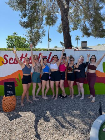 Nine friends in colorful activewear raising glasses of bubbly and smiling in front of a bright mural on a sunny Southwest backyard with palm trees and gravel ground.