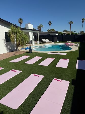 Sunny suburban backyard with a kidney-shaped pool, palm trees and lounge chairs; pink yoga mats lined up on green artificial turf for an outdoor fitness class.