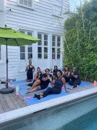 Seven women smiling on blue yoga mats beside a backyard pool, enjoying a poolside yoga/fitness session on a wooden deck in front of a white clapboard house with bamboo and a green umbrella, some holding drinks.