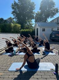 Group outdoor yoga class on blue mats in a sunny suburban driveway, participants in seated side-stretch poses near a white shed and trees.