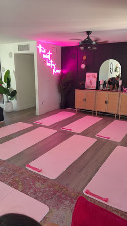 Boutique workout studio with rows of pink exercise mats on wood floors, neon pink wall sign casting a rosy glow, rattan sideboard, mirror and potted plants — ready for a group fitness class.