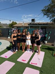 Six women in activewear posing on pink mats for an outdoor backyard yoga / group fitness session by a pool, colorful floral mural on a cinderblock wall under a sunny blue sky