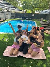 Five friends cheering with drinks and handheld fans at a sunny backyard pool party on artificial grass with loungers, umbrellas and colorful floaties.