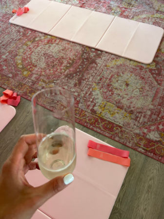 Hand with white manicure holding a champagne flute over a home workout setup: pink folding exercise mats and coral resistance bands on a faded patterned rug