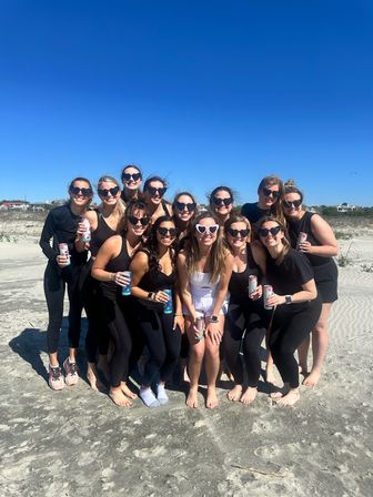 Smiling group of friends posing on a sunny sandy beach with clear blue sky, most wearing black activewear and sunglasses and one in white, holding canned drinks for a casual coastal celebration.