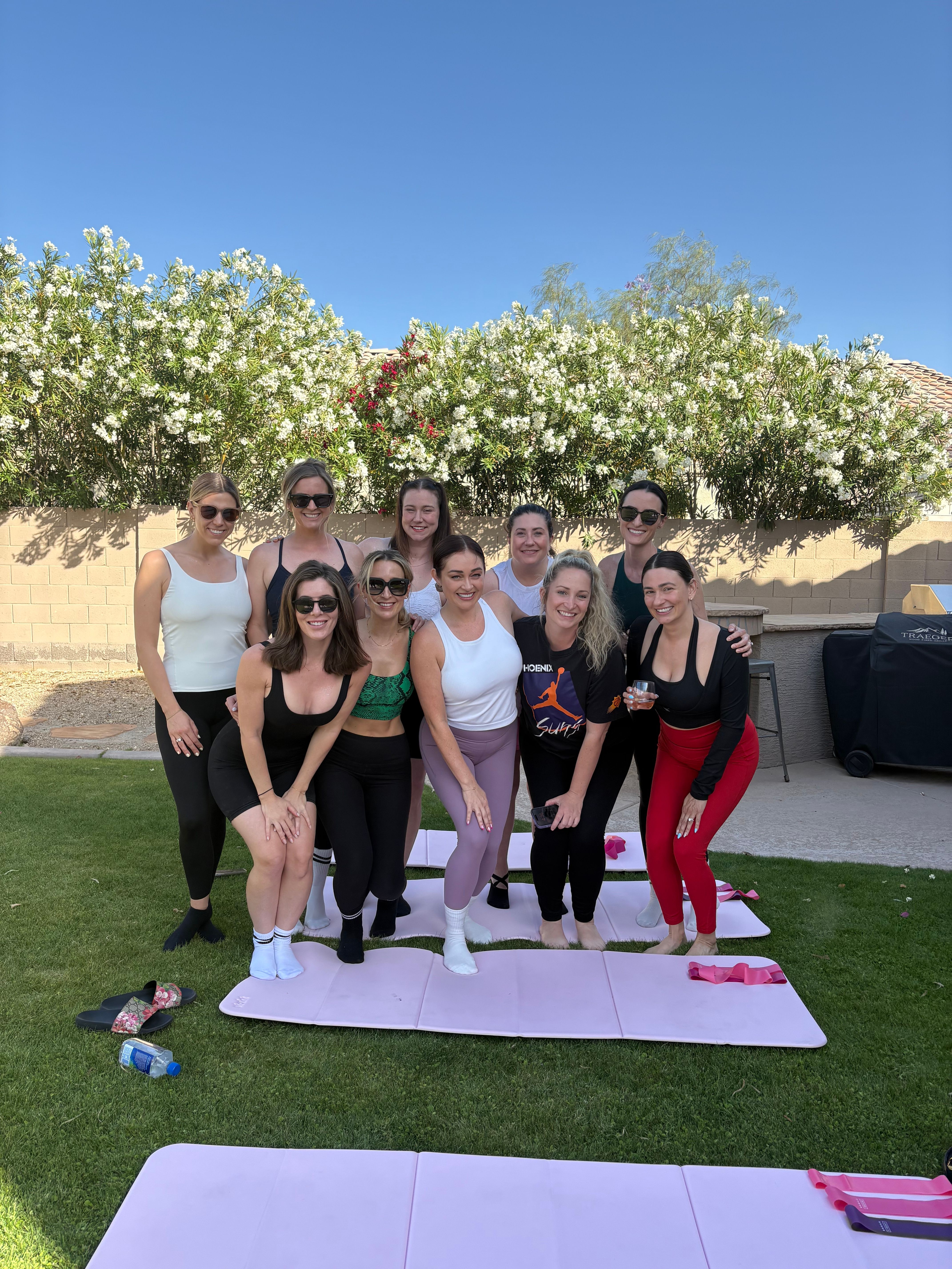 Ten women in colorful activewear smiling and posing on pink yoga mats in a sunny suburban backyard with flowering hedges and a clear blue sky.
