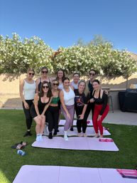 Ten women in colorful activewear smiling and posing on pink yoga mats in a sunny suburban backyard with flowering hedges and a clear blue sky.