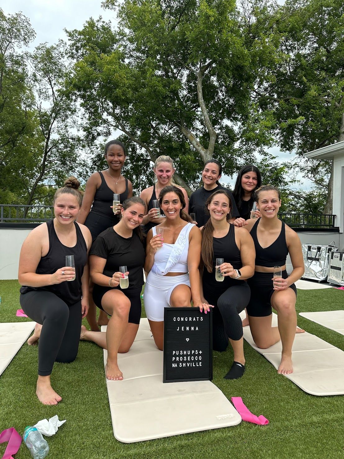 Rooftop fitness celebration in Nashville: nine women in activewear pose on outdoor yoga mats, smiling and toasting with prosecco beside a congratulatory letter board, with trees in the background.