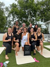 Rooftop fitness celebration in Nashville: nine women in activewear pose on outdoor yoga mats, smiling and toasting with prosecco beside a congratulatory letter board, with trees in the background.